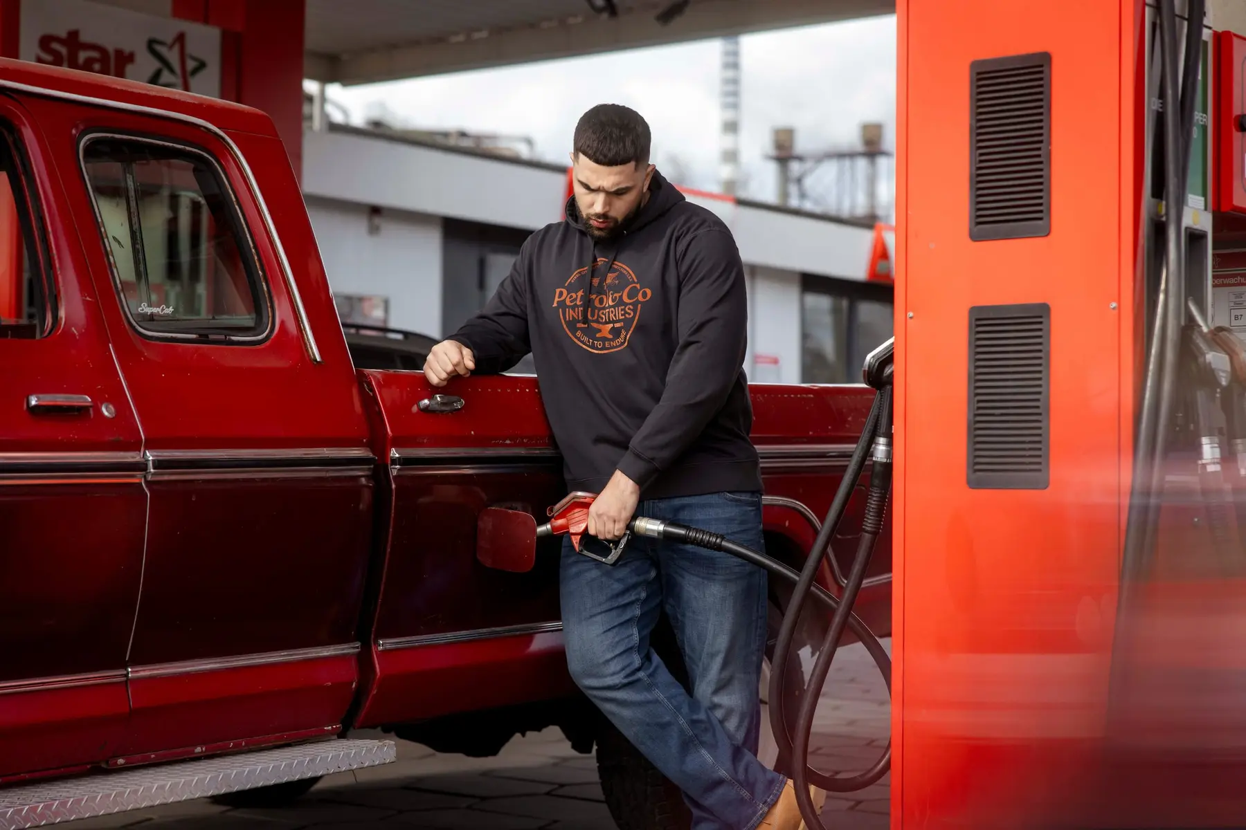 Mann in Sweatshirt und Jeans an einer Zapfsäule neben einem roten Pick-up