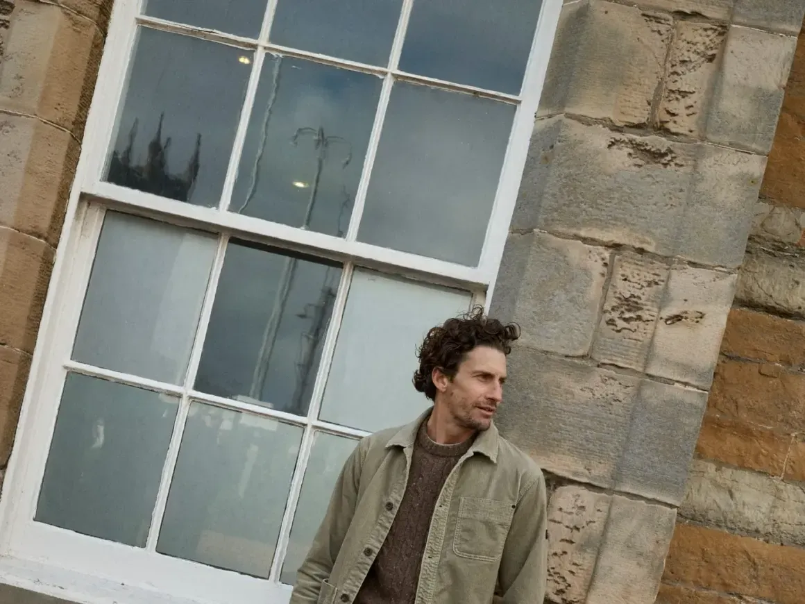 Mann in olivfarbener Jacke und heller Hose vor einer Steinhauswand